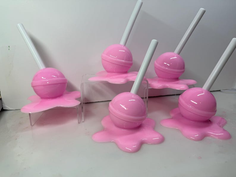 Op de afbeelding: Roze lolly's met witte stokjes, rustend op roze, gesmolten uitziende bases. De lolly's zijn gerangschikt op heldere acrylstandaards, wat een speelse en grillige esthetiek cre&euml;ert. Snoepgoed-thema tegen een witte achtergrond.