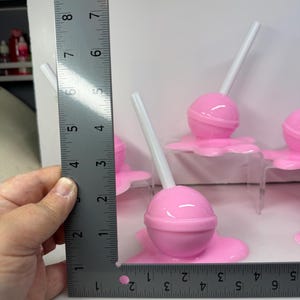 Op de afbeelding: Verschillende roze lolly's met witte stokjes. De lolly's staan op een roze basis. De liniaal geeft aan dat de lolly's ongeveer 5 cm hoog zijn.