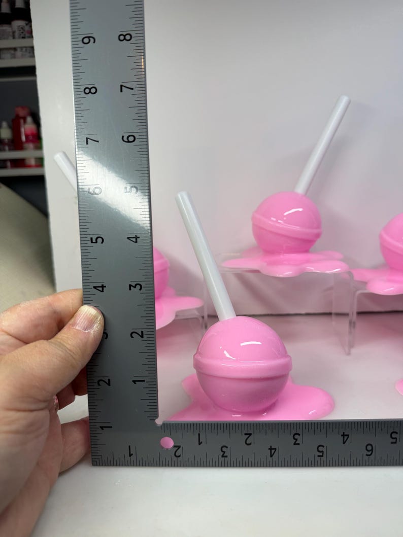Op de afbeelding: Verschillende roze lolly's met witte stokjes. De lolly's staan op een roze basis. De liniaal geeft aan dat de lolly's ongeveer 5 cm hoog zijn.