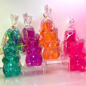 May include: A collection of colorful gummy bear candies in shades of green, purple, orange, and pink. Each bear is individually wrapped in a clear cellophane bag with a twist tie. The bears are displayed on clear acrylic stands.