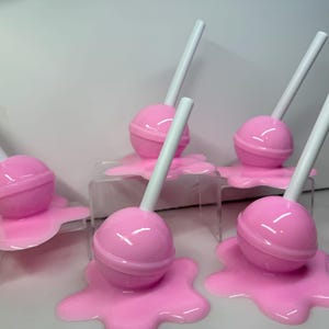 Op de afbeelding: Zes roze lolly's met witte stokjes, die elk lijken te smelten op een bijpassend roze oppervlak. De lolly's zijn gerangschikt op kleine, transparante displaystandaards, waardoor een speelse, snoep-thema presentatie ontstaat.