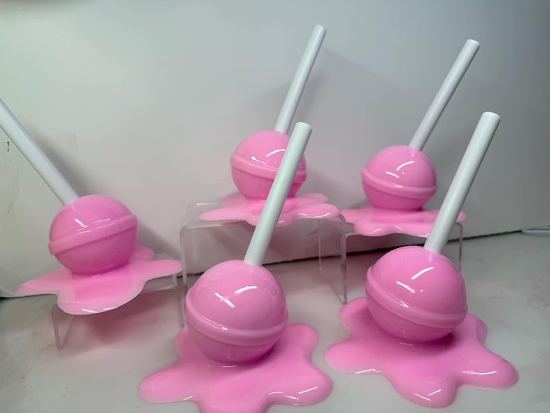 Op de afbeelding: Zes roze lolly's met witte stokjes, die elk lijken te smelten op een bijpassend roze oppervlak. De lolly's zijn gerangschikt op kleine, transparante displaystandaards, waardoor een speelse, snoep-thema presentatie ontstaat.