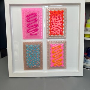 May include: A white framed artwork featuring four colorful, rectangular designs resembling frosted pastries. Each pastry has a different color frosting and sprinkles, including pink, red, blue, and orange. The artwork is displayed on a white background.