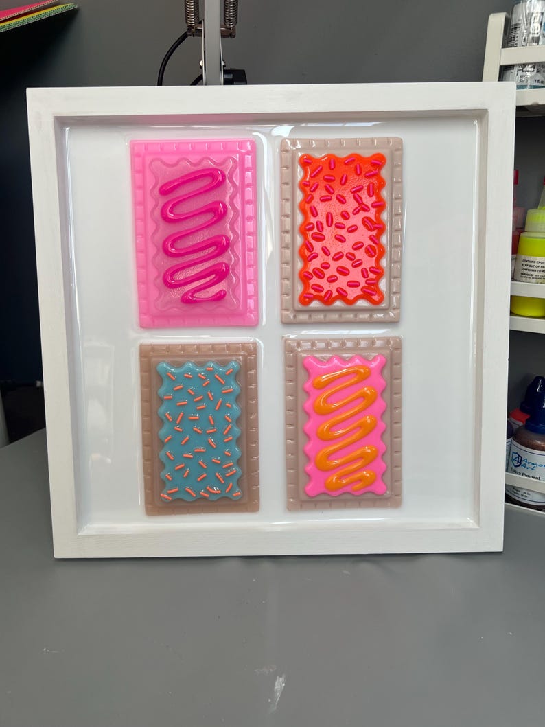 May include: A white framed artwork featuring four colorful, rectangular designs resembling frosted pastries. Each pastry has a different color frosting and sprinkles, including pink, red, blue, and orange. The artwork is displayed on a white background.