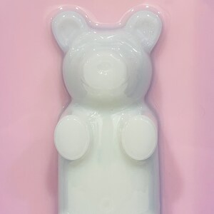 Puede incluir: Un molde blanco y transl&uacute;cido con forma de oso de goma sobre un fondo rosa claro s&oacute;lido. La figura del oso presenta orejas redondeadas, una superficie lisa y extremidades definidas, creando una silueta clara y elevada sobre la superficie plana.