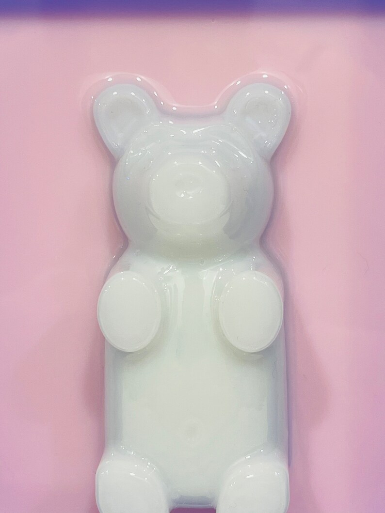 Puede incluir: Un molde blanco y transl&uacute;cido con forma de oso de goma sobre un fondo rosa claro s&oacute;lido. La figura del oso presenta orejas redondeadas, una superficie lisa y extremidades definidas, creando una silueta clara y elevada sobre la superficie plana.