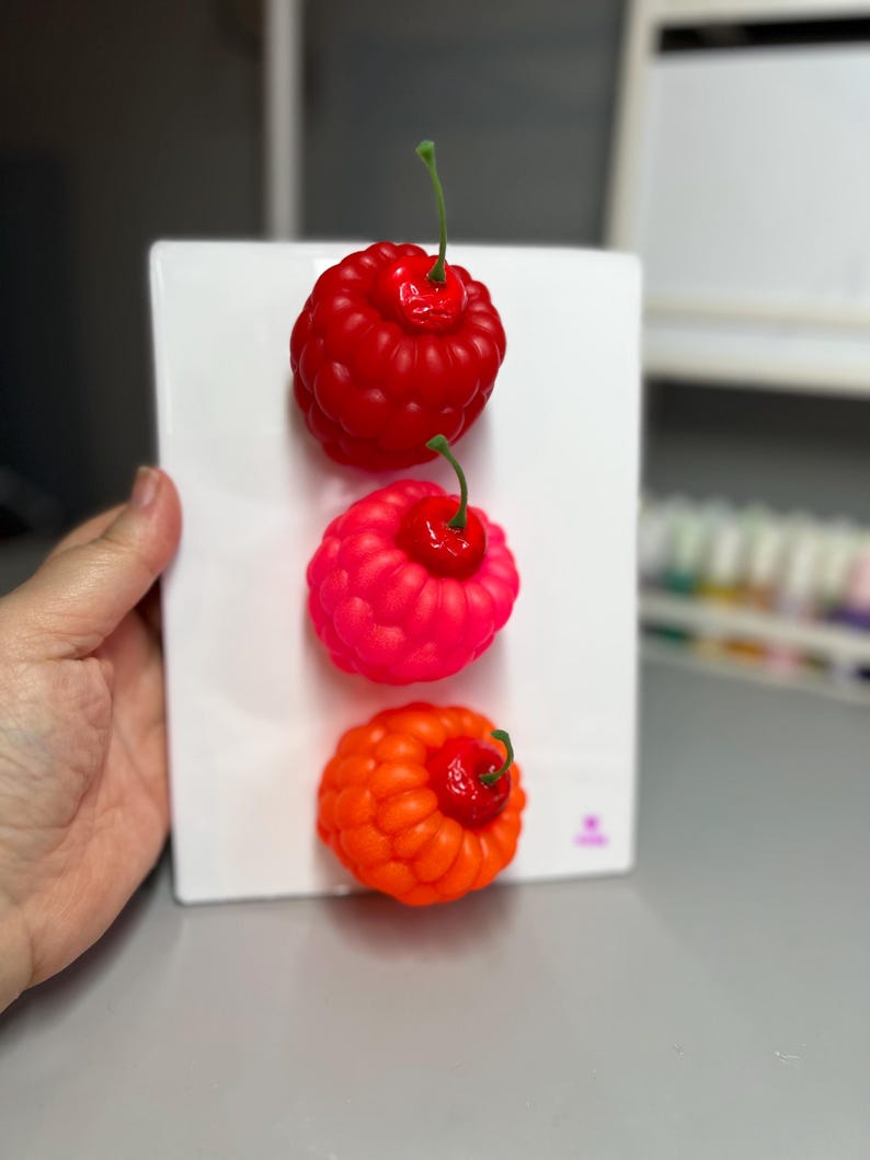 May include: Three colorful, artificial cherries in red, pink, and orange are displayed on a white rectangular surface. Each cherry has a green stem and a textured surface, creating a vibrant and playful arrangement.