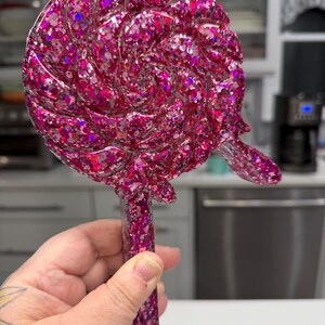 May include: A lollipop-shaped object made of resin with a vibrant pink and magenta glitter finish. The circular head features a swirling, textured pattern resembling a classic candy, while the straight handle is also filled with matching glitter.