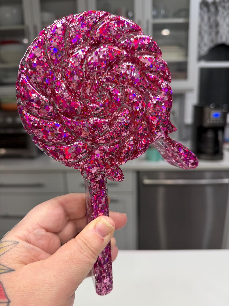 May include: A lollipop-shaped object made of resin with a vibrant pink and magenta glitter finish. The circular head features a swirling, textured pattern resembling a classic candy, while the straight handle is also filled with matching glitter.