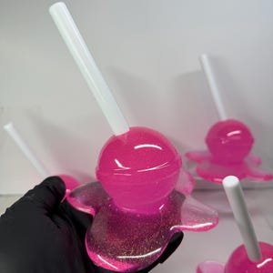 Puede incluir: Primer plano de varias piruletas rosas de resina con palitos blancos. Las piruletas tienen una base brillante en forma de flor. El color rosa es vibrante y la resina es brillante. El fondo es blanco liso.