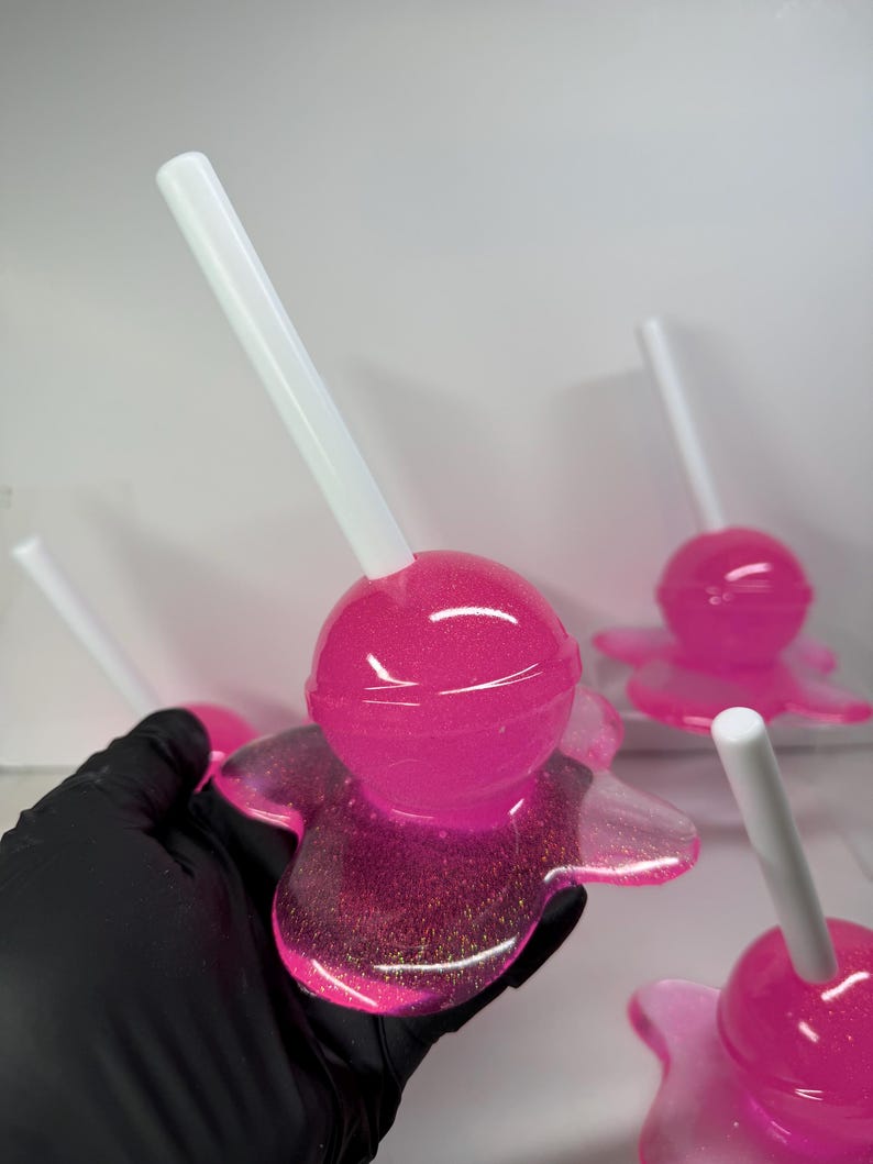 Puede incluir: Primer plano de varias piruletas rosas de resina con palitos blancos. Las piruletas tienen una base brillante en forma de flor. El color rosa es vibrante y la resina es brillante. El fondo es blanco liso.