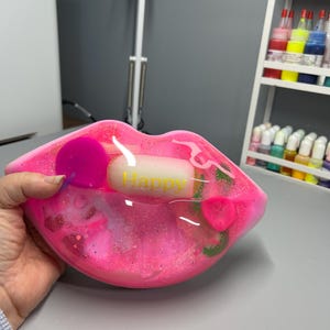 Peut inclure: Un plat à bijoux en forme de lèvres en résine rose avec des paillettes et une petite barre blanche qui dit "Happy" à l'intérieur.