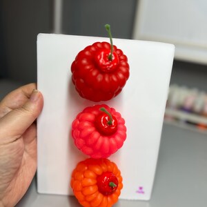 May include: Three colorful, decorative berry-shaped objects are displayed on a white surface. The top berry is a deep red, the middle one is a vibrant pink-red, and the bottom one is a bright orange. Each berry has a green stem.