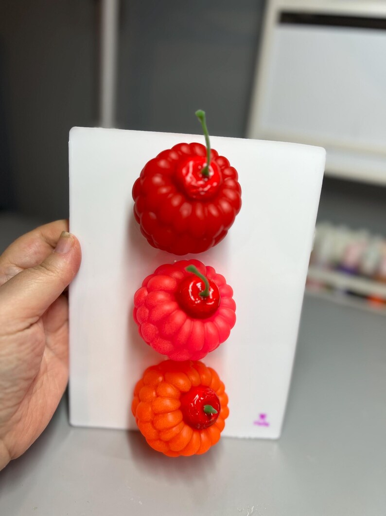 May include: Three colorful, decorative berry-shaped objects are displayed on a white surface. The top berry is a deep red, the middle one is a vibrant pink-red, and the bottom one is a bright orange. Each berry has a green stem.