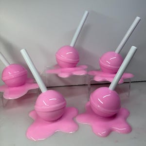 Op de afbeelding: Een verzameling roze lolly's met witte stokjes, elk geplaatst op een bijpassende roze, gesmolten uitziende basis. De lolly's zijn gerangschikt op heldere, verhoogde platforms, wat een speelse en grillige presentatie cre&euml;ert.