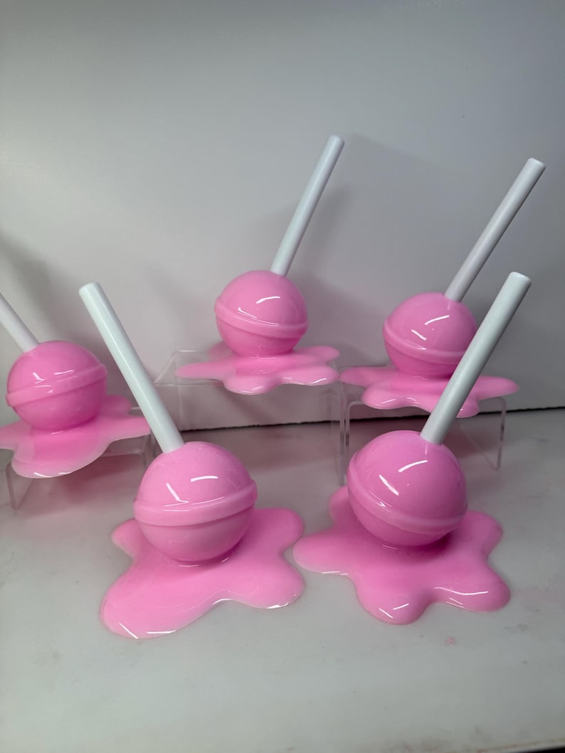 Op de afbeelding: Een verzameling roze lolly's met witte stokjes, elk geplaatst op een bijpassende roze, gesmolten uitziende basis. De lolly's zijn gerangschikt op heldere, verhoogde platforms, wat een speelse en grillige presentatie cre&euml;ert.