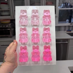 Pu&ograve; includere: Uno stampo rettangolare trasparente riempito con dodici orsetti gommosi rosa. Gli orsetti gommosi sono disposti in tre file da quattro. Lo stampo &egrave; tenuto davanti a uno sfondo di cucina.