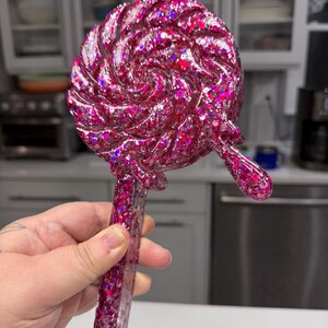 May include: A handheld mirror shaped like a round lollipop with a swirl pattern. The object is made of a translucent pink resin filled with chunky holographic glitter that reflects light. It features a long, straight handle and a small drip.