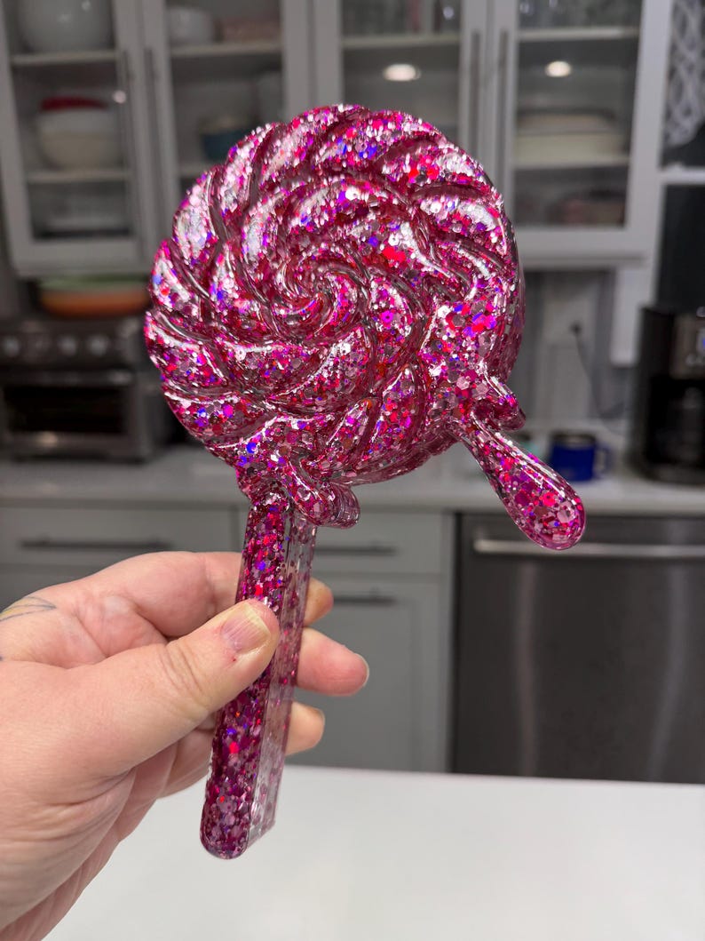 May include: A handheld mirror shaped like a round lollipop with a swirl pattern. The object is made of a translucent pink resin filled with chunky holographic glitter that reflects light. It features a long, straight handle and a small drip.