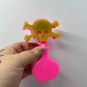 Op de afbeelding: Een felroze flesstopper met een gele schedel en kruisbotten ontwerp. De schedel heeft een gele tot groene gradiënt. De stopper wordt getoond tegen een witte achtergrond.