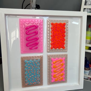 May include: A framed artwork featuring four colorful, stylized Pop-Tarts. Each pastry has a different frosting and sprinkle design: pink with pink frosting, orange with orange sprinkles, blue with orange sprinkles, and pink with orange frosting. The frame is white.