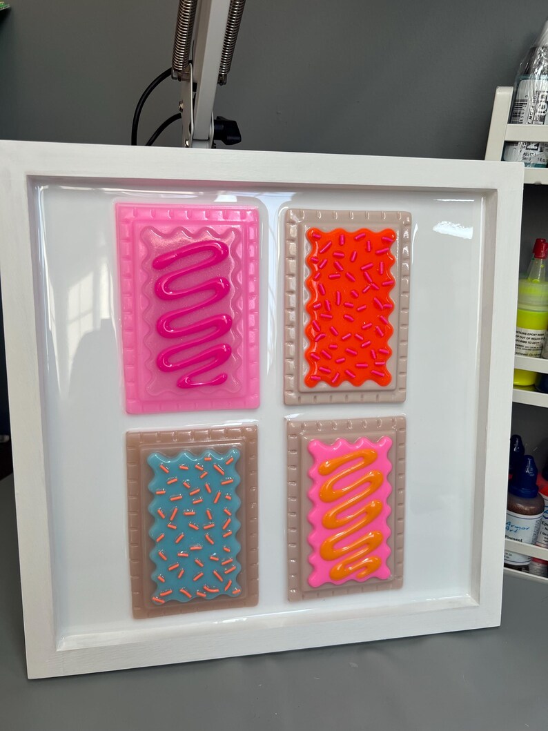 May include: A framed artwork featuring four colorful, stylized Pop-Tarts. Each pastry has a different frosting and sprinkle design: pink with pink frosting, orange with orange sprinkles, blue with orange sprinkles, and pink with orange frosting. The frame is white.