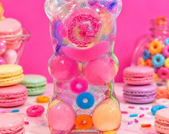 May include: A clear resin bear figurine filled with colourful embedded items including pink sprinkles, small pink and blue ring-shaped sweets, and larger round pink and orange spheres, creating a textured, multi-coloured decorative object.