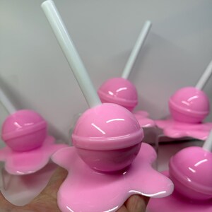 Op de afbeelding: Roze lolly's met witte stokjes, die lijken te smelten op een roze ondergrond. De lolly's hebben een glanzende afwerking en de afbeelding is een close-up, die de details van het snoepgoed benadrukt.