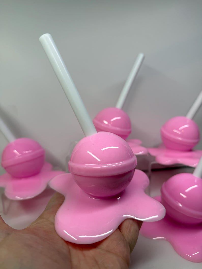 Op de afbeelding: Roze lolly's met witte stokjes, die lijken te smelten op een roze ondergrond. De lolly's hebben een glanzende afwerking en de afbeelding is een close-up, die de details van het snoepgoed benadrukt.