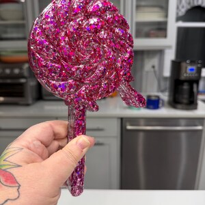 May include: A lollipop-shaped object made of translucent pink resin filled with various sizes of holographic pink and silver glitter. The circular head features a swirling, textured pattern, and it includes a straight handle at the bottom.