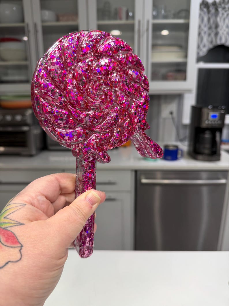 May include: A lollipop-shaped object made of translucent pink resin filled with various sizes of holographic pink and silver glitter. The circular head features a swirling, textured pattern, and it includes a straight handle at the bottom.