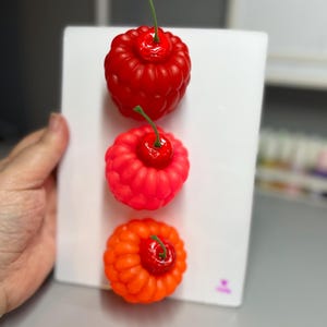 May include: Three colorful, stylized cherry sculptures. The top cherry is red, the middle is a lighter red, and the bottom is orange. Each has a green stem and sits on a white surface. The cherries are arranged vertically.