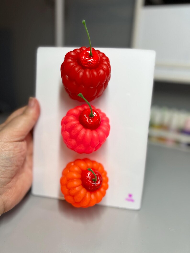 May include: Three colorful, stylized cherry sculptures. The top cherry is red, the middle is a lighter red, and the bottom is orange. Each has a green stem and sits on a white surface. The cherries are arranged vertically.