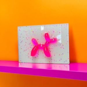 May include: A rectangular, white resin art piece featuring a bright pink balloon dog design. The resin is speckled with colourful glitter. The artwork is displayed on a bright pink shelf against an orange background.