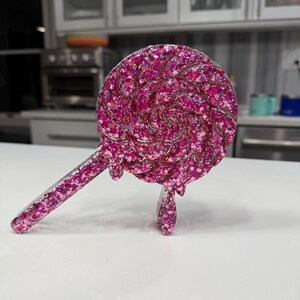 May include: A circular lollipop-shaped object made of translucent pink resin filled with dense pink glitter. The design features a swirling pattern on the circular head with a straight handle extending to the left and a small drip detail at the bottom.