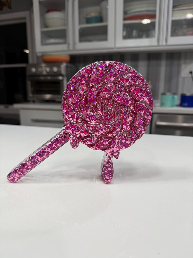 May include: A circular lollipop-shaped object made of translucent pink resin filled with dense pink glitter. The design features a swirling pattern on the circular head with a straight handle extending to the left and a small drip detail at the bottom.
