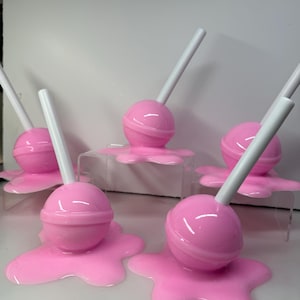 Op de afbeelding: Een verzameling roze lolly's met witte stokjes, die lijken te smelten op een oppervlak. De lolly's hebben een glanzende roze kleur en het gesmolten effect voegt een grillige toets toe. De afbeelding is goed belicht en benadrukt de details.
