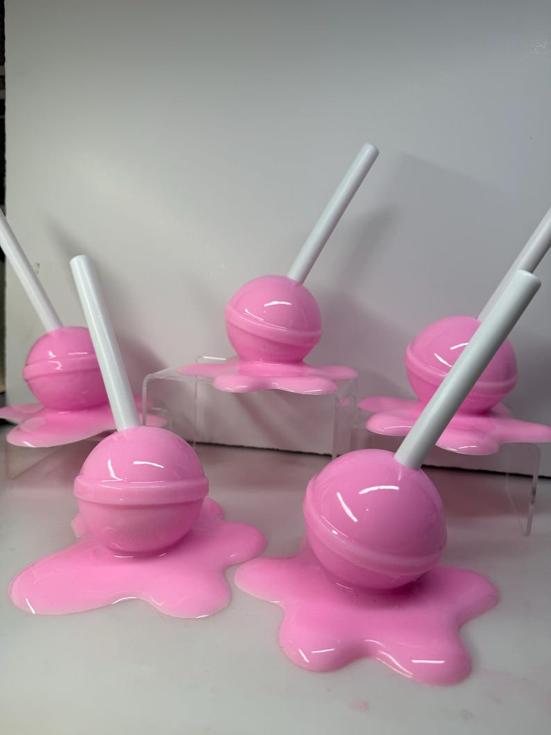 Op de afbeelding: Een verzameling roze lolly's met witte stokjes, die lijken te smelten op een oppervlak. De lolly's hebben een glanzende roze kleur en het gesmolten effect voegt een grillige toets toe. De afbeelding is goed belicht en benadrukt de details.