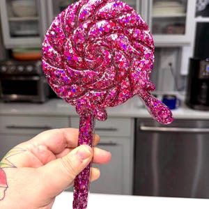 May include: A circular lollipop-shaped object made of pink resin filled with dense, multi-sized pink and magenta glitter. The surface features a raised spiral pattern and a small, rounded drip detail on the right side, attached to a straight handle.