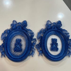Op de afbeelding: Twee blauwe glitter fotolijstjes met witte binnenkanten en een blauwe glitter teddybeer in elk lijstje. De lijstjes zijn versierd met blauwe en witte linten.
