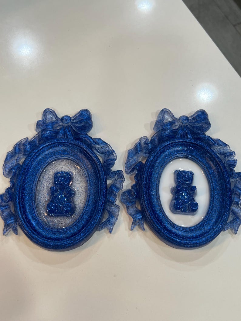Op de afbeelding: Twee blauwe glitter fotolijstjes met witte binnenkanten en een blauwe glitter teddybeer in elk lijstje. De lijstjes zijn versierd met blauwe en witte linten.