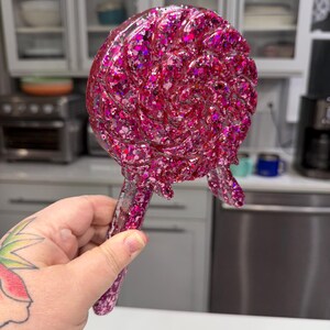 May include: A lollipop-shaped decorative object crafted from clear resin filled with vibrant pink and magenta glitter. The circular head features a swirling pattern with a textured surface, and it includes a straight handle extending from the base.