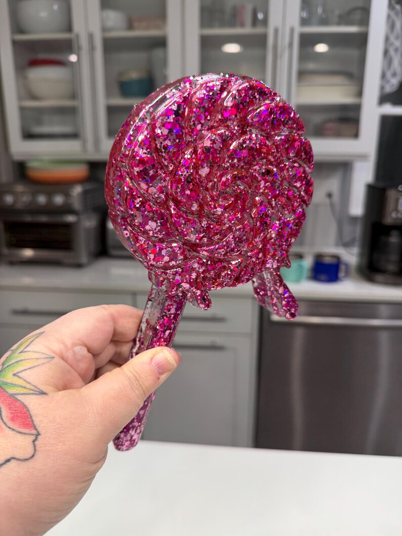 May include: A lollipop-shaped decorative object crafted from clear resin filled with vibrant pink and magenta glitter. The circular head features a swirling pattern with a textured surface, and it includes a straight handle extending from the base.