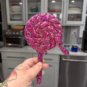 May include: A circular lollipop-shaped object featuring a swirling pattern and a small drip detail on the side. The entire piece is crafted from a translucent pink resin material embedded with numerous reflective pink and purple glitter flakes.