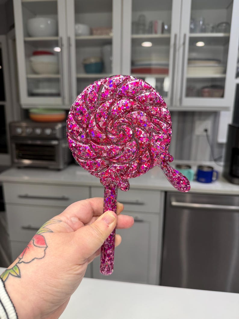 May include: A circular lollipop-shaped object featuring a swirling pattern and a small drip detail on the side. The entire piece is crafted from a translucent pink resin material embedded with numerous reflective pink and purple glitter flakes.