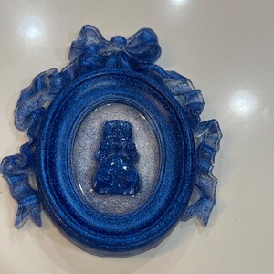 Op de afbeelding: Een blauw glitterhars ornament met een wit lint en strik. Het ornament heeft een blauw glitterhars beeldje van een persoon in een blauw glitterhars ovaal frame.