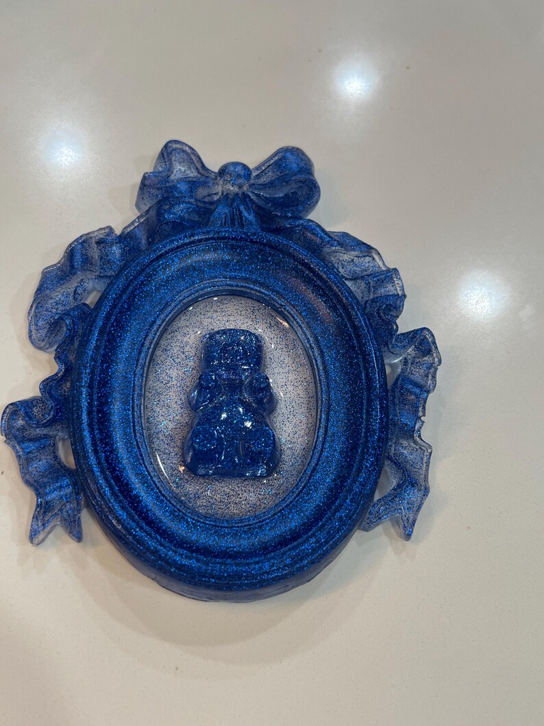 Op de afbeelding: Een blauw glitterhars ornament met een wit lint en strik. Het ornament heeft een blauw glitterhars beeldje van een persoon in een blauw glitterhars ovaal frame.