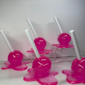 Puede incluir: Seis piruletas de color rosa brillante con palitos blancos, que parecen derretirse sobre una superficie blanca. Las piruletas tienen un acabado brillante y est&aacute;n dispuestas de forma l&uacute;dica. El efecto de caramelo derretido a&ntilde;ade un toque caprichoso.