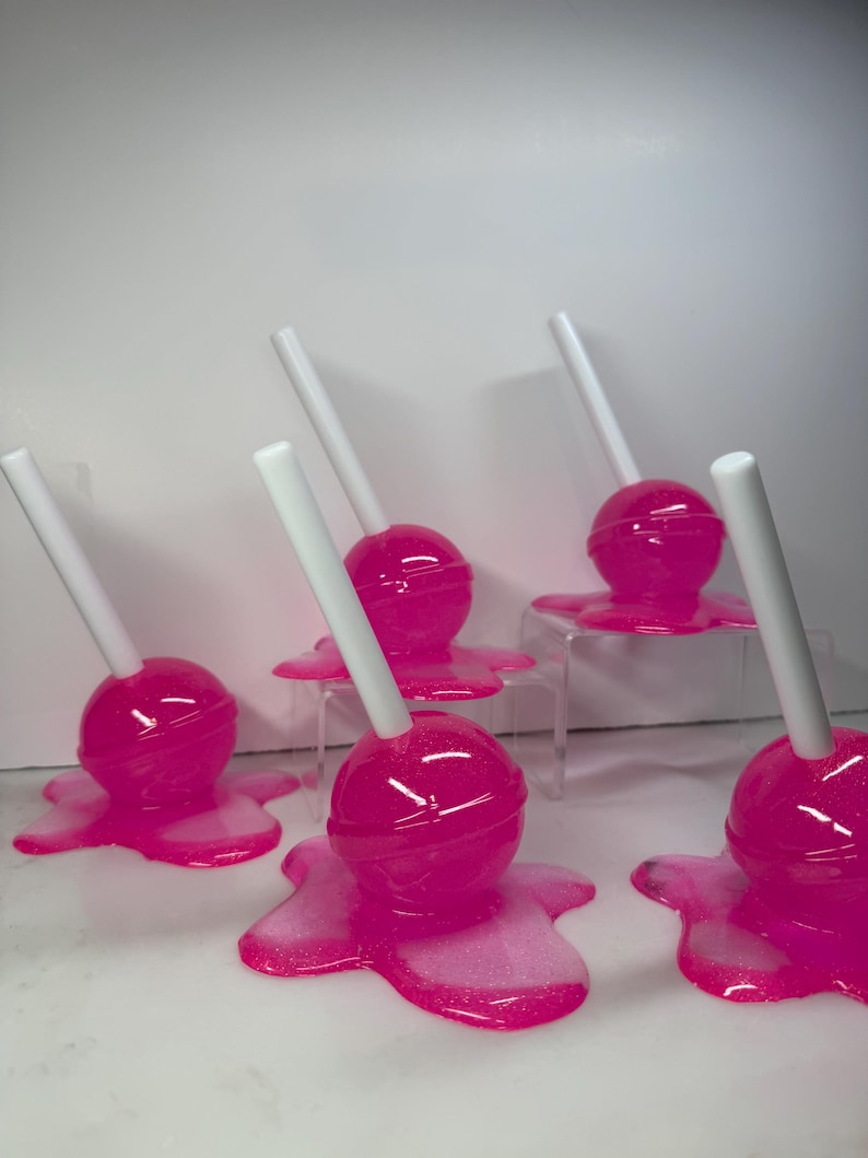 Puede incluir: Seis piruletas de color rosa brillante con palitos blancos, que parecen derretirse sobre una superficie blanca. Las piruletas tienen un acabado brillante y est&aacute;n dispuestas de forma l&uacute;dica. El efecto de caramelo derretido a&ntilde;ade un toque caprichoso.