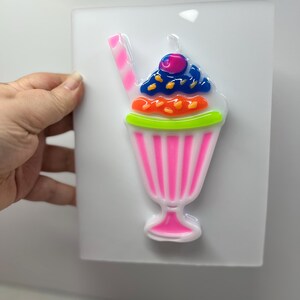 May include: A colorful, cartoon-style milkshake design. The milkshake features a pink glass with vertical stripes, topped with blue, orange, and green elements, and a cherry. A pink and white striped straw is included.